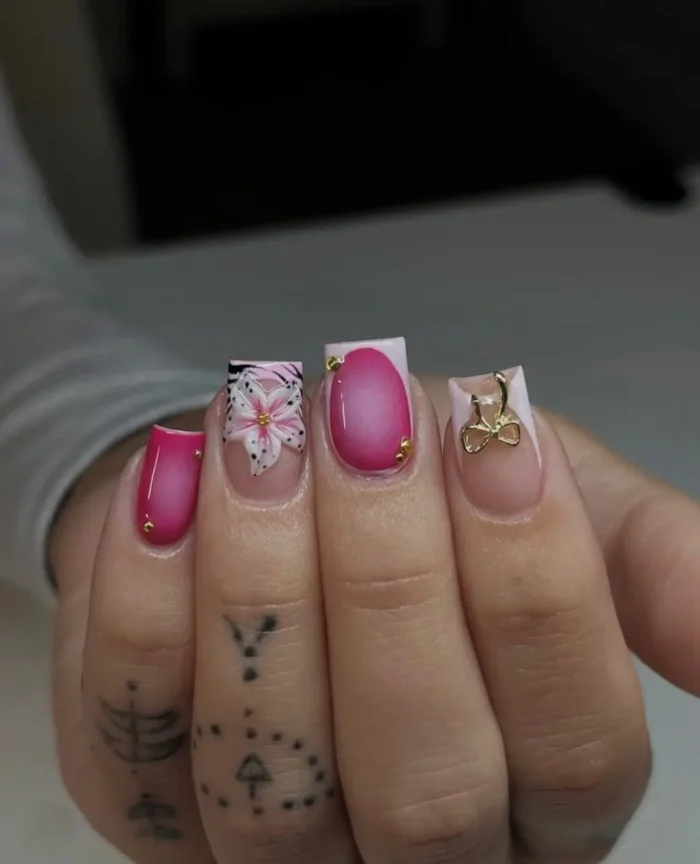 Short square nails showcasing a playful mix: vibrant pink nails with gold stud accents, a graphic pink and white floral on a light tip, and a classic French tip with a gold bow charm.