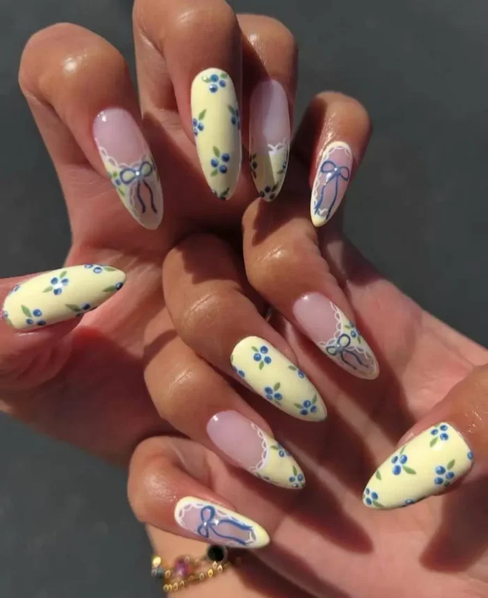 A charming manicure with butter yellow nails adorned with blue berry art, and accent nails with blue bows and lace details.