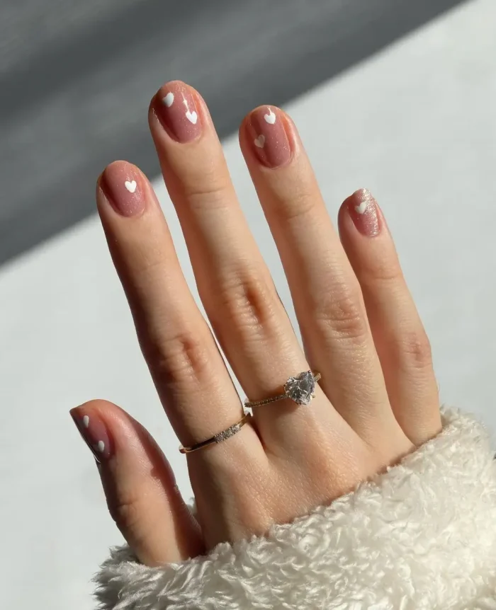 Short rounded nails with a glossy rosy nude polish featuring small white heart accents painted near the cuticles.