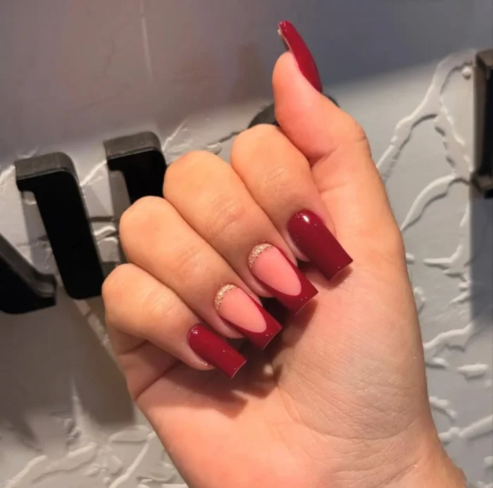A sophisticated deep red manicure with a French tip accent nail featuring a delicate gold glitter lunula detail.