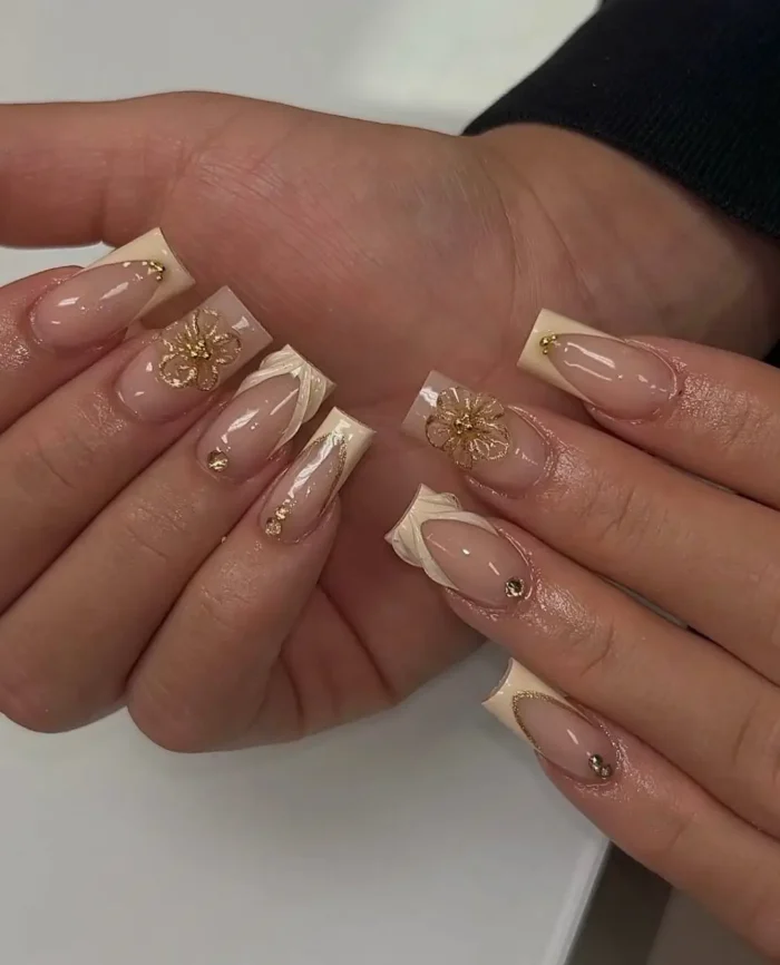 Long square nails with luxurious creamy off-white French tips, adorned with intricate 3D gold floral charms, sculpted draped effects, and delicate gold stud and gem embellishments.