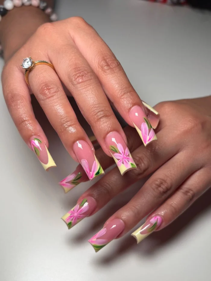 Very long square nails with pale yellow French tips, adorned with hand-painted pink flowers and green leaves extending onto the sheer pink nail bed.