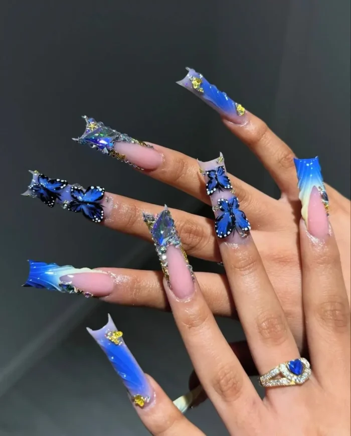 Fantastical long sculpted nails with vibrant blue butterfly-wing tips, 3D blue butterflies, and gold foil details.