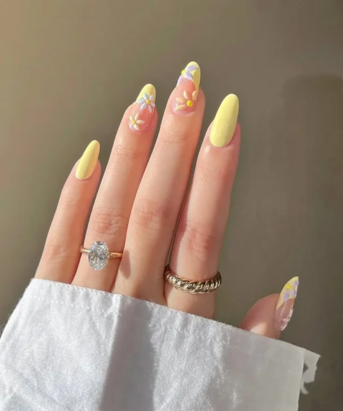 A hand with almond nails wearing a butter yellow manicure, with two accent nails featuring sweet daisy floral art.
