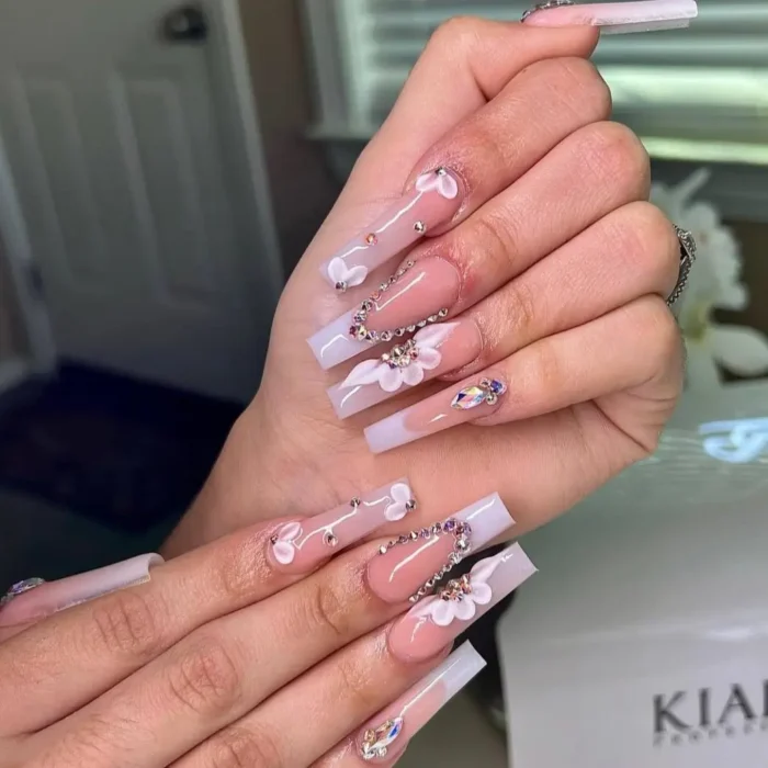 Long square nails featuring 3D white flower embellishments and sparkling iridescent rhinestone accents.