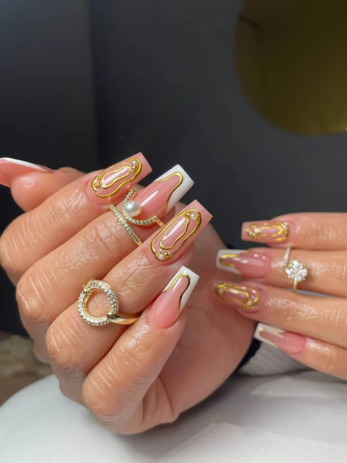 Long square nails showcasing a classic white French manicure dramatically enhanced with 3D abstract gold metallic designs and small pearl embellishments.