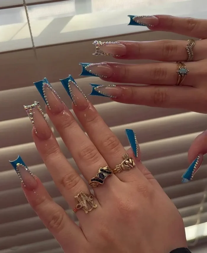 Extra-long clear sculpted nails showcasing a vibrant blue V-shaped French tip, lavishly outlined and adorned with dense rows of clear, sparkling rhinestones.