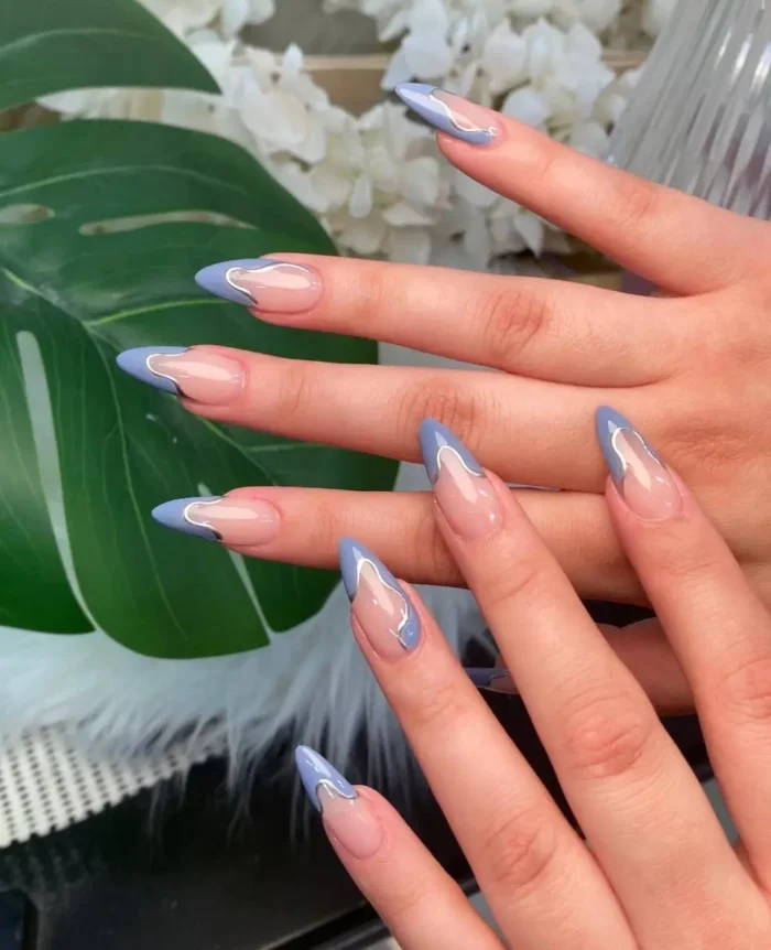 Long almond nails with a modern French design featuring wavy periwinkle blue tips outlined in silver on a sheer base.