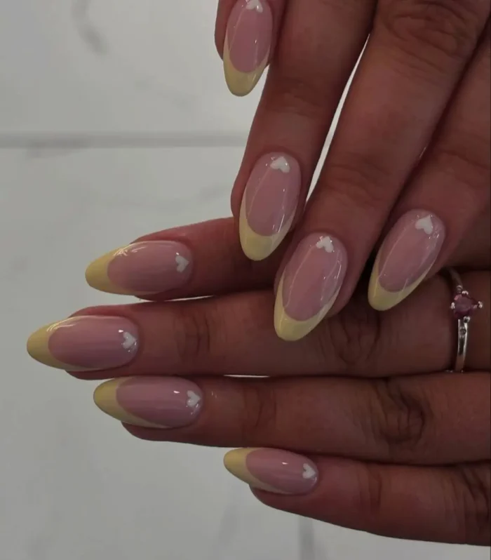 Butter yellow French manicure on almond nails with a small white heart accent at the base of each nail.
