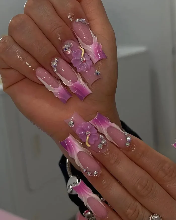 Long sculpted nails with purple and pink flame-style French tips, 3D purple flowers, and rhinestone embellishments.