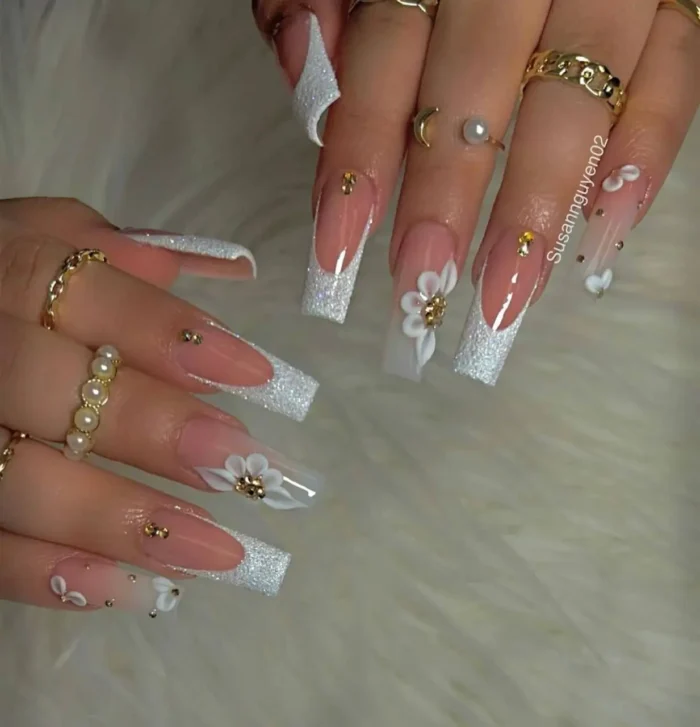 Long square nails with intensely sparkling white and silver glitter French tips, accented with 3D white flowers and gold studs.