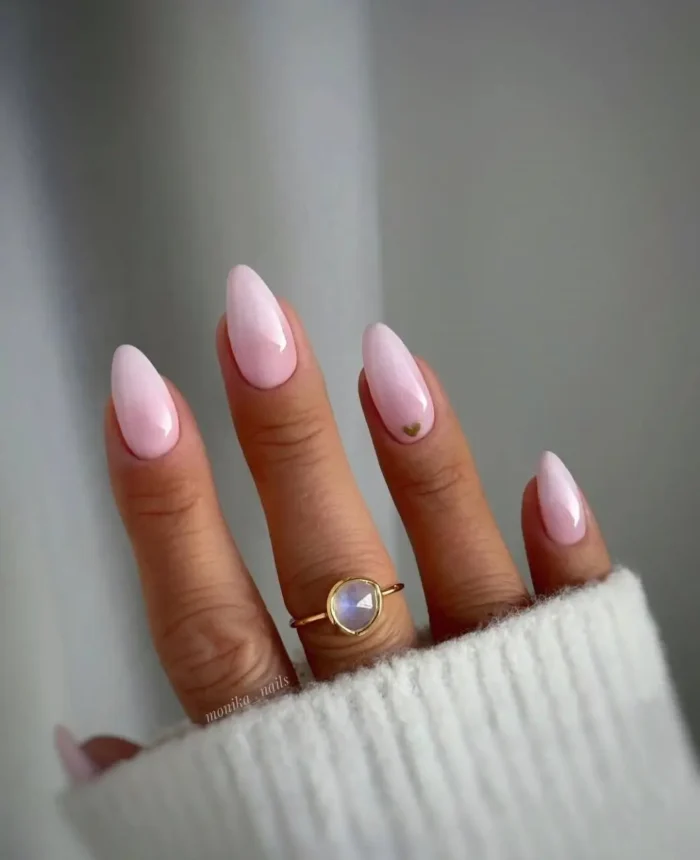 Long almond-shaped nails in glossy baby pink, subtly enhanced with a tiny, delicate gold heart accent near the cuticle on one nail.