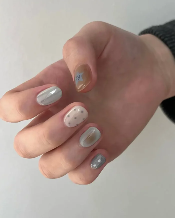 Short rounded nails with an abstract mixed design featuring muted blue and beige color washes, polka dots, star accents, and a swirl symbol.