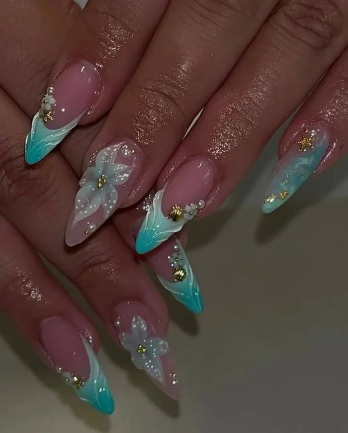 Vibrant turquoise ocean-themed summer nail art on long stiletto nails, with 3D white flowers, seashells, and gold details.
