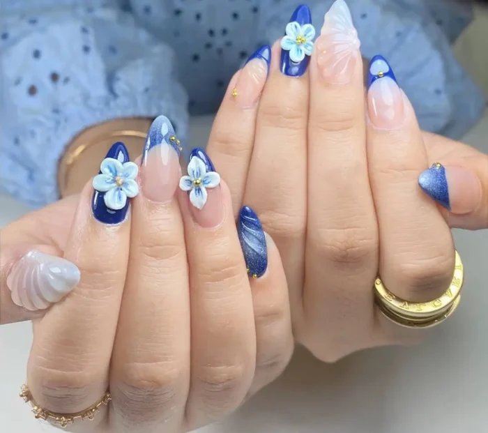 Long almond nails with mixed designs in royal blue and white, including 3D flowers, cat-eye tips, marble effects, and gold stud details.