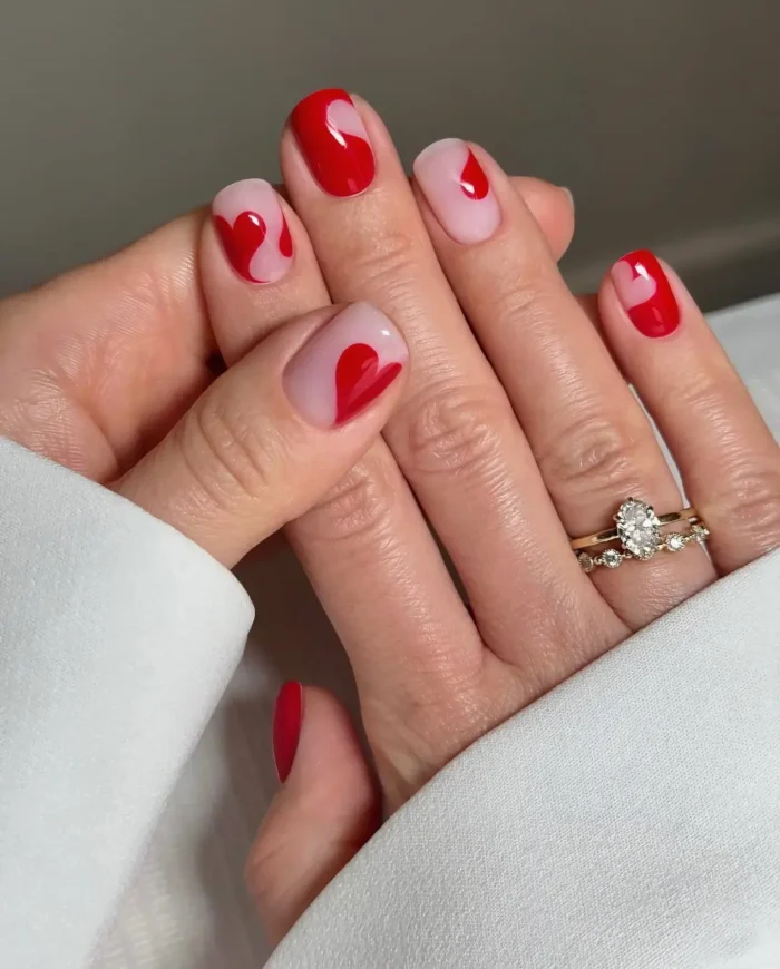 Short square nails with a sheer nude base featuring abstract red heart shapes created using negative space, plus a solid red nail.