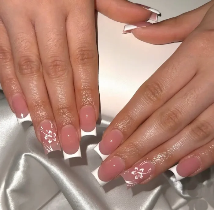Elegant French tip nails with a soft pink base and crisp white tips, enhanced by intricate white floral designs.