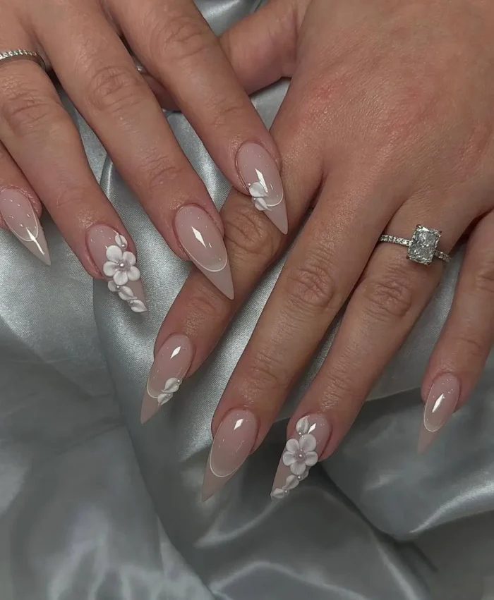 Long stiletto nails with a sheer milky nude base, featuring delicate white 3D flower accents and a subtle white micro French outline at the tips.