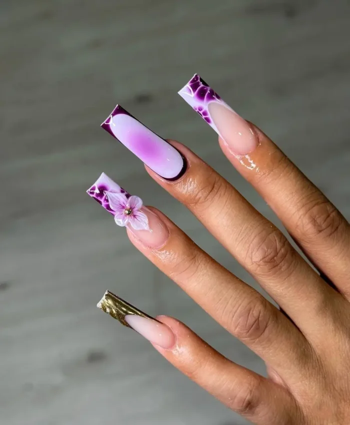 Long square nails with mixed designs: purple reptilian print V-cut French tips, a purple aura nail, a 3D lavender flower, and a textured gold tip.
