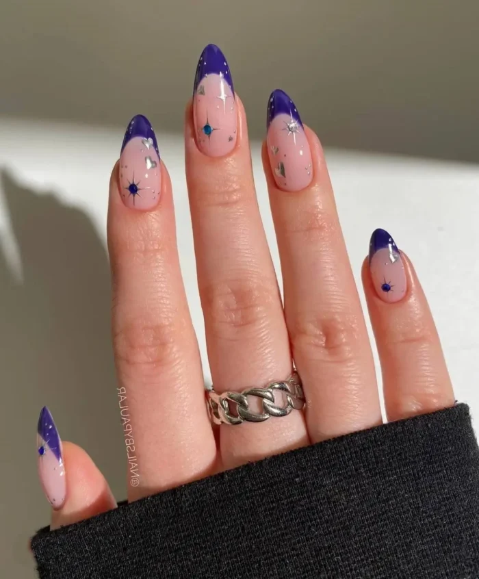Almond nails with a glittery sheer pink base, deep indigo V-shaped French tips, and small starburst accents with rhinestones.