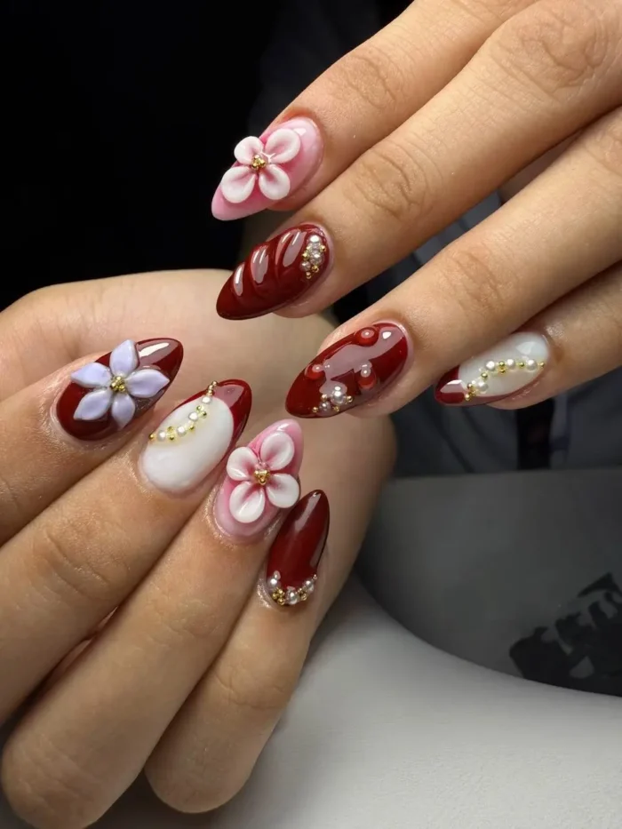 An elegant red nail design with 3D pink and lavender floral charms, pearl accents, and glossy red nails.