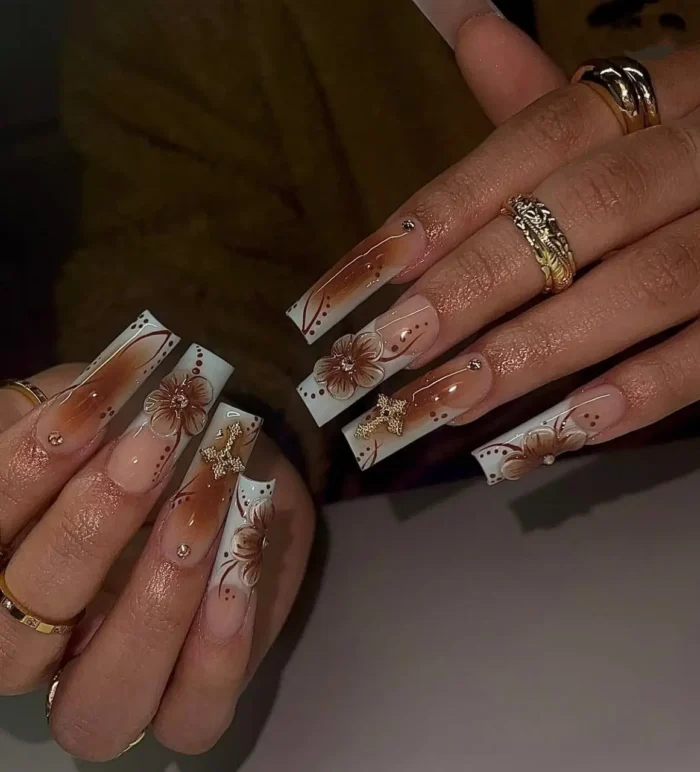 Long square nails in shades of brown featuring ombré, French tips with dot accents, 3D brown flowers, and gold butterfly charms.