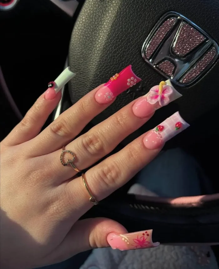 Playful long square nails with a summer garden theme, showcasing a mix of designs including a prominent 3D hibiscus flower, painted red strawberries on a checkered tip, and a tiny ladybug charm.