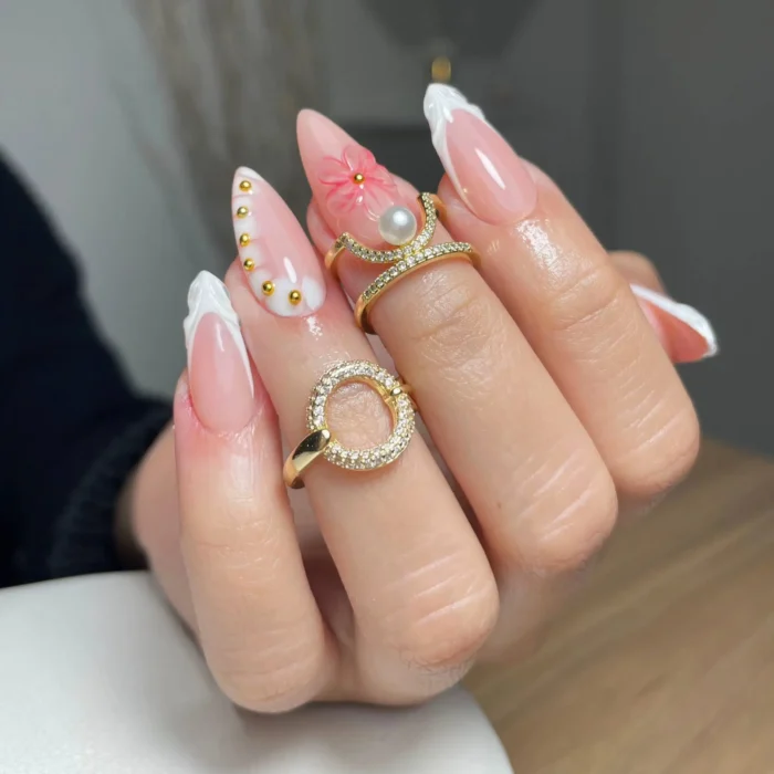 Long stiletto nails with textured white French tips, featuring a 3D pink flower with a large pearl center, and gold stud accents on a sheer pink base.