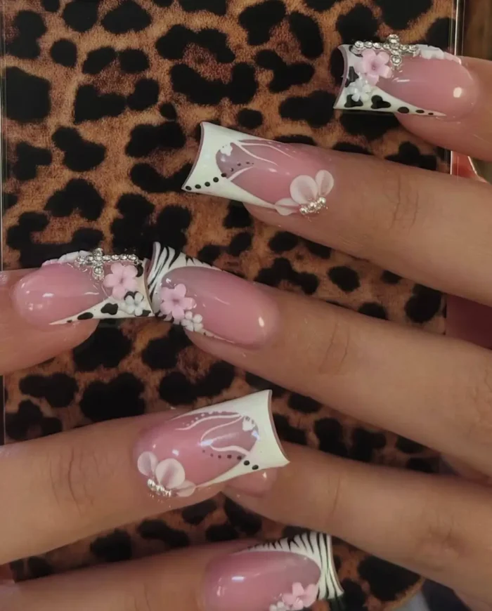 Long sculpted nails showcasing a French manicure with mixed animal prints like cow and zebra, playfully adorned with 3D pink and white floral embellishments and rhinestone details.