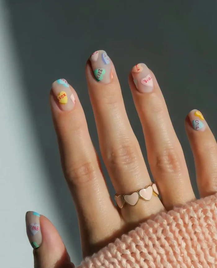 Short rounded nails with a sheer nude base decorated with tiny pastel conversation heart candy designs featuring red text.