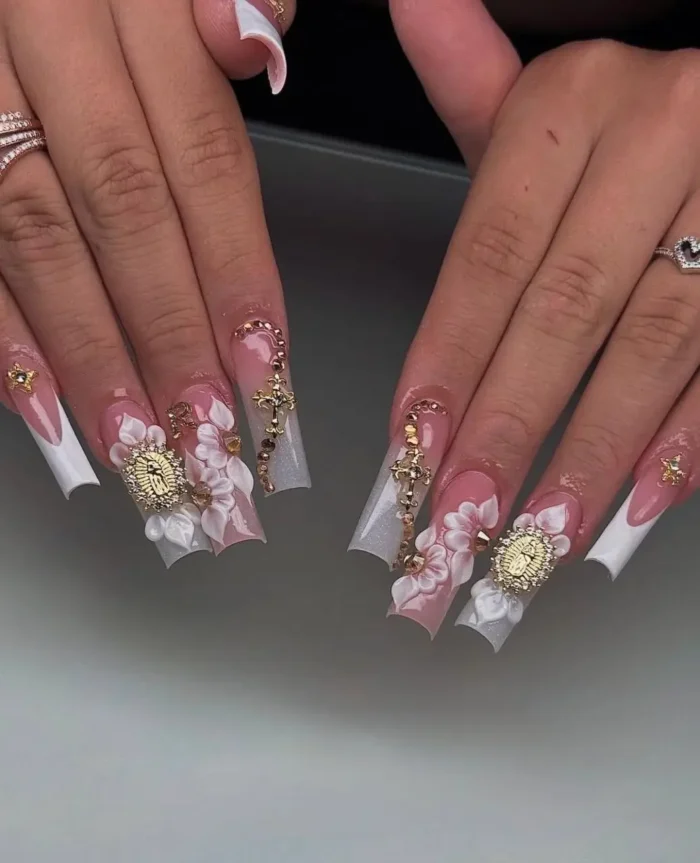 Long coffin-shaped nails with white French tips, adorned with 3D white and pink flowers, gold religious icon charms, and gold cross charms.