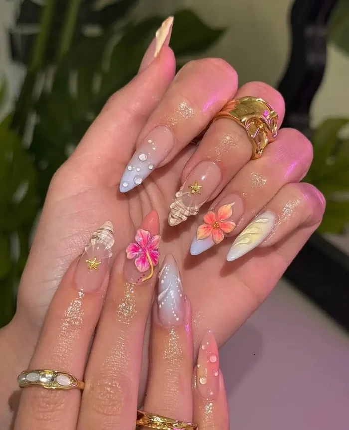 Long stiletto nails with a whimsical summer design, mixing 3D seashells, flowers, pastel tips, plaid, and gold accents.