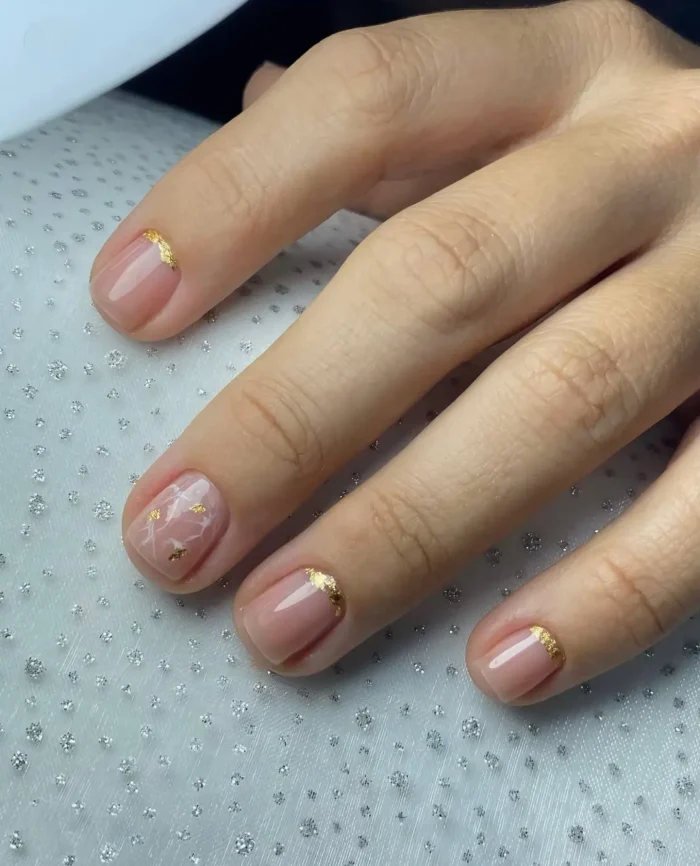 Short square nails with a sheer pink base featuring gold foil accents near the cuticles and a white marble effect with gold foil on one nail.