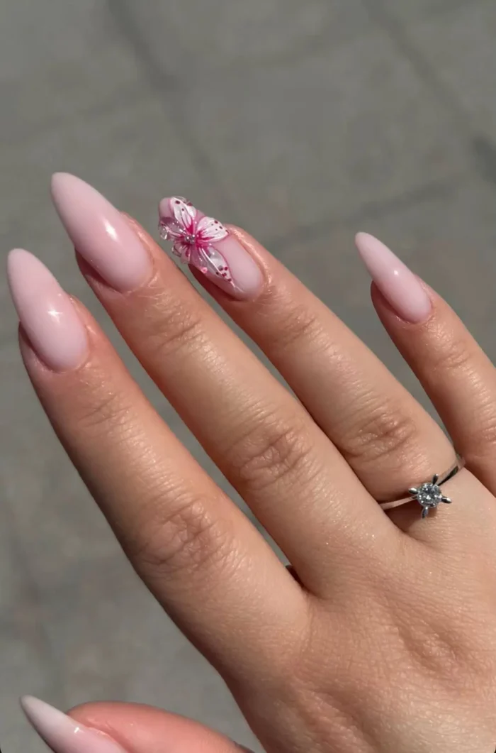 Long almond-shaped nails with a sheer milky pink base, featuring a single accent nail with a detailed hand-painted or 3D pink and white flower with a jeweled center.