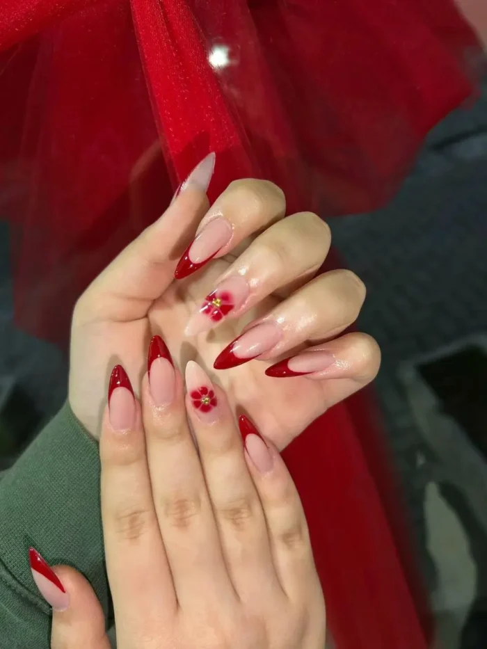 An elegant red French tip manicure on almond nails with 3D red floral accents and gold bead centers.