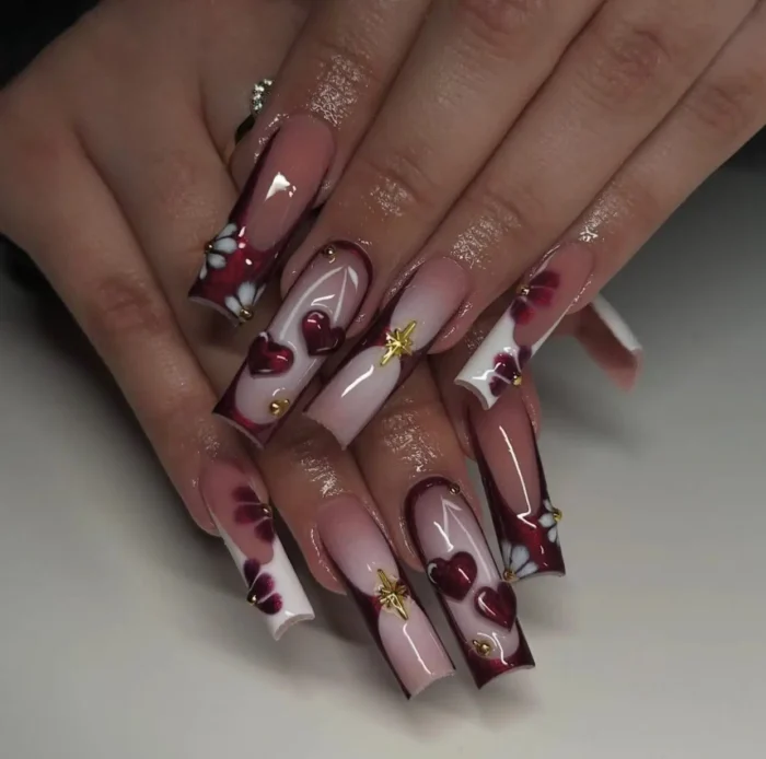 Long square nails with a white to pink ombré, adorned with deep maroon 3D hearts, maroon and white floral tip designs, and gold studs.
