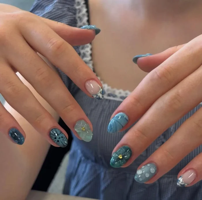 Short oval nails with mixed designs in shades of blue and teal, including abstract patterns, 3D flowers, lace tips, dots, and textured finishes.