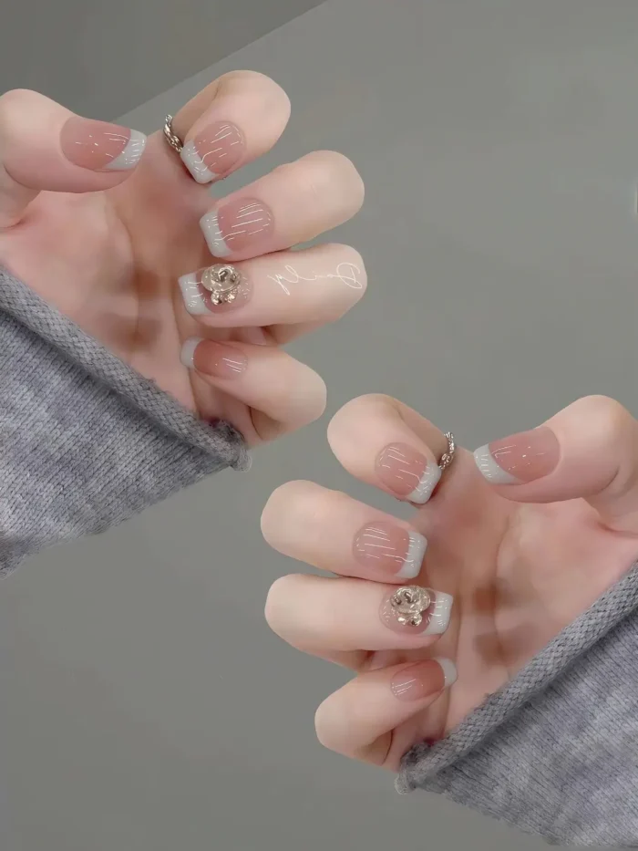 Short square nails with a sheer nude base, sage green French tips, subtle white line details, and a 3D gold rose charm accent.