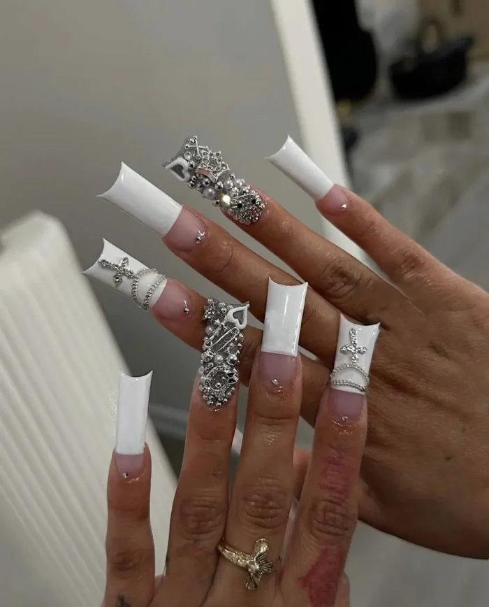 Statement long square nails with white French tips, lavishly adorned with an assortment of silver embellishments including cross charms, chains, chrome hearts, and dense stud work.