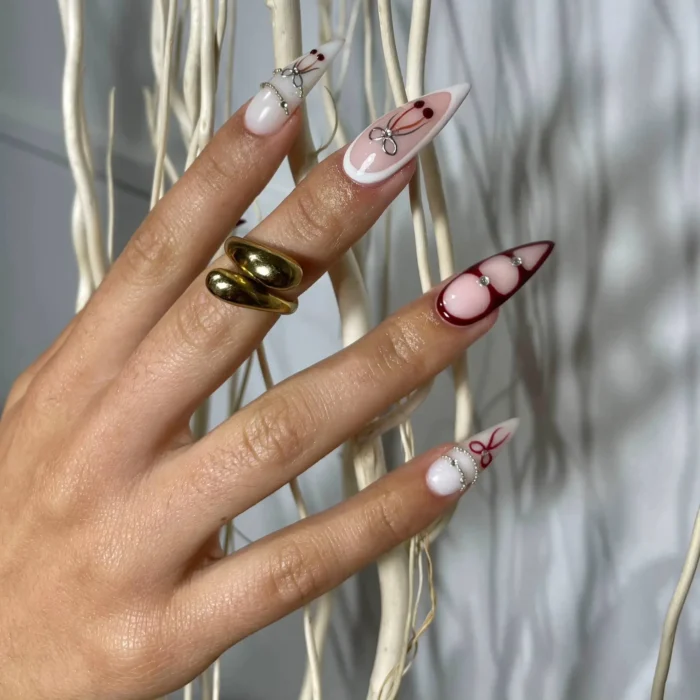 Long stiletto nails featuring a mix of designs including deep burgundy negative space outlines, hand-painted bow motifs, and fine silver chain and crystal accents.