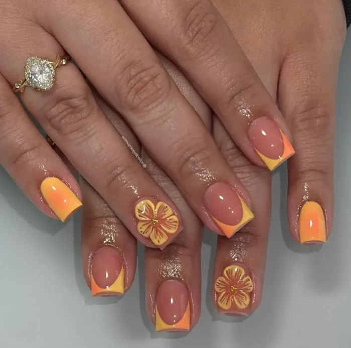 Short to medium square nails displaying a two-tone ombré French tip in shades of orange and yellow, with some nails adorned with a matching 3D tropical flower.