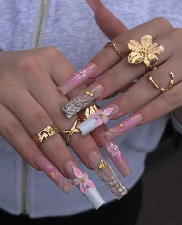 Extra-long nails with a mix of pink floral designs, marbled wave patterns, seashell charms, and gold stud embellishments.