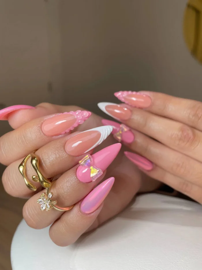 Long stiletto nails in a vibrant pink theme, featuring solid pink nails with a large iridescent bow charm, textured pink 'sugar bead' details, and white V-French tips.