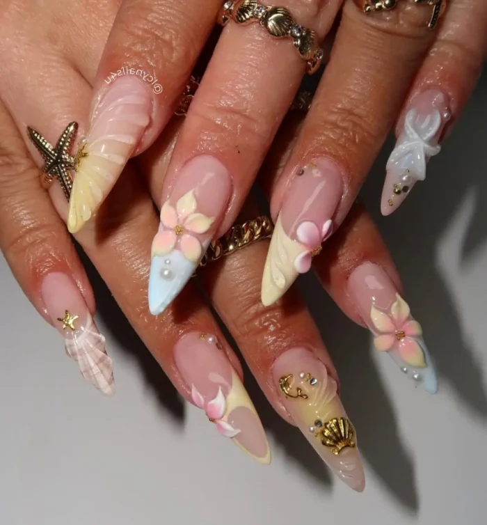 Long stiletto vacation nails with an intricate seaside theme, showcasing detailed 3D seashells, starfish, pastel flowers, and gold accents.