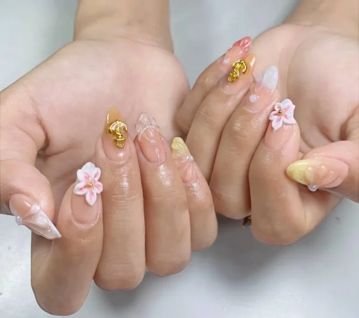 Long stiletto nails with mixed designs featuring sheer and ombre bases adorned with 3D sculpted flowers, chunky gold charms, and pearl accents.