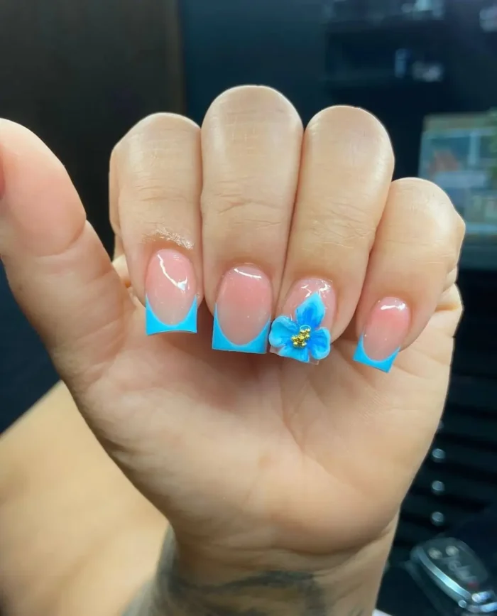 Short to medium square nails with bright sky blue French tips on a shimmery pink base, with one nail elegantly adorned with a matching 3D blue flower with gold bead details.