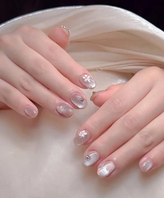 Rounded nails with pearly silver cat-eye base polish, featuring delicate white painted florals, swirls, and silver stud accents.