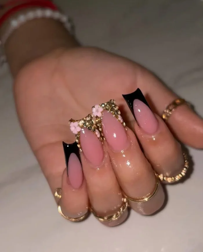 Almond-shaped nails with a sharp black V-shaped French manicure on a sheer pink base, highlighted by an accent nail with 3D pink flowers and dense gold crystal embellishments at the cuticle.