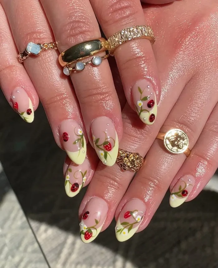 A detailed manicure featuring butter yellow French tips decorated with intricate strawberry, vine, and ladybug nail art.