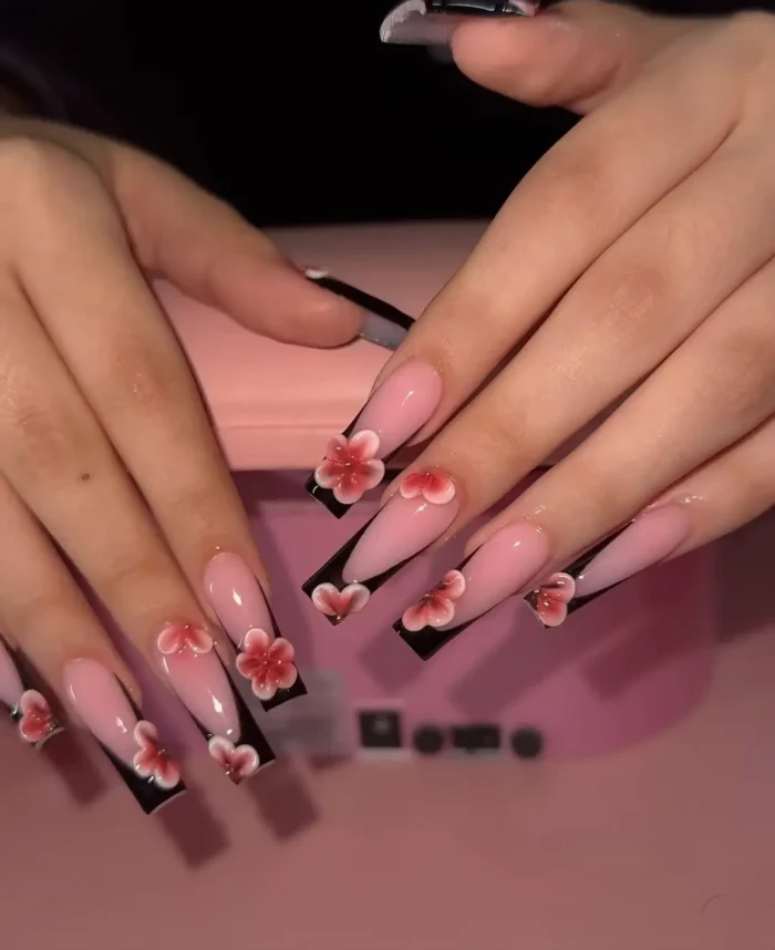 Long coffin-shaped nails featuring a black V-French tip design and 3D sculpted pink flowers on a sheer pink base.